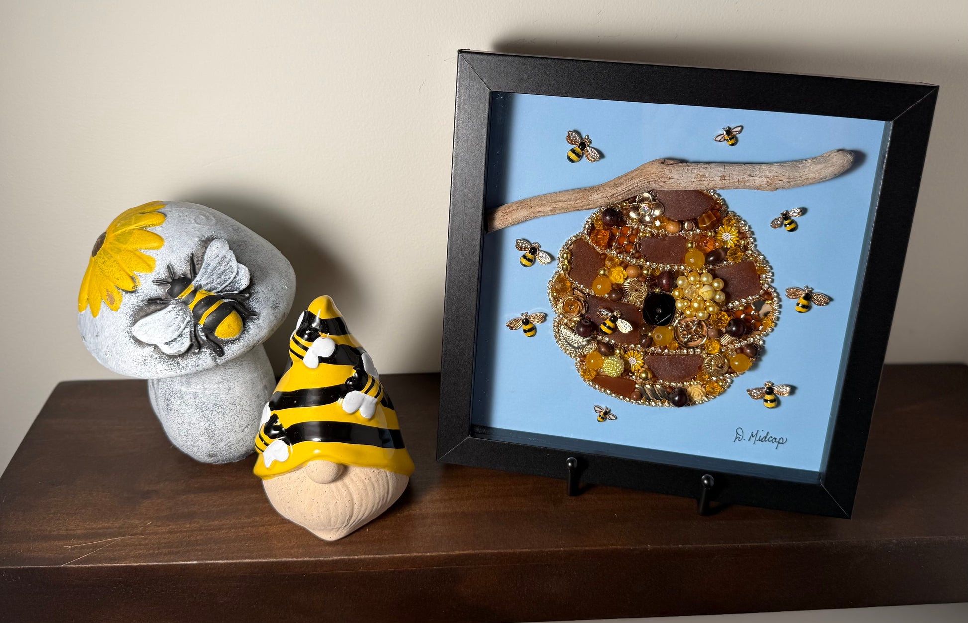 Decorative items including a framed artwork of a sea glass beehive, a mushroom with a bee design, and a gnome with a bee hat on a wooden surface.