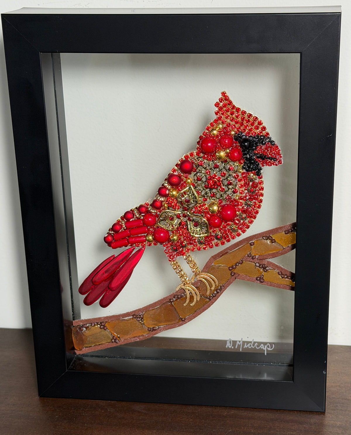 A framed shadow box art piece featuring a red and gold beaded cardinal perched on a brown branch, set inside a black frame and displayed on a dark wooden surface against a white wall.