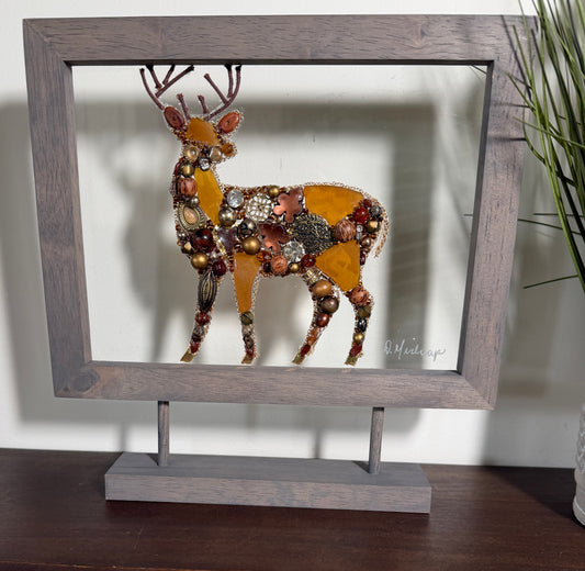 A framed deer wall art piece displayed on a stand. The deer silhouette is filled with amber-colored panels and many small rhinestones and beads, set inside a rustic gray wooden frame with a matching base.