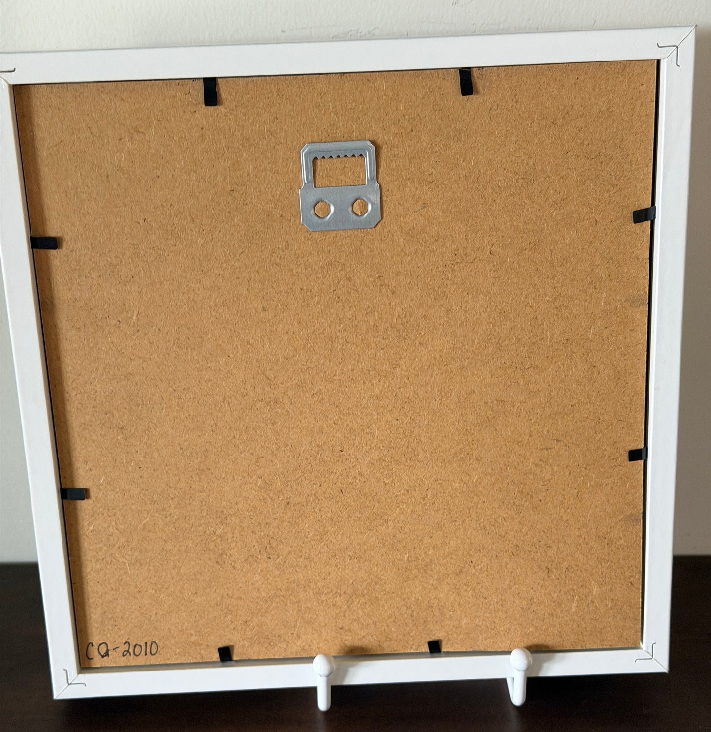 The back of a square picture frame with a white border, a brown cork backing board, a silver metal hanging loop, and white feet, shown standing on a dark surface.