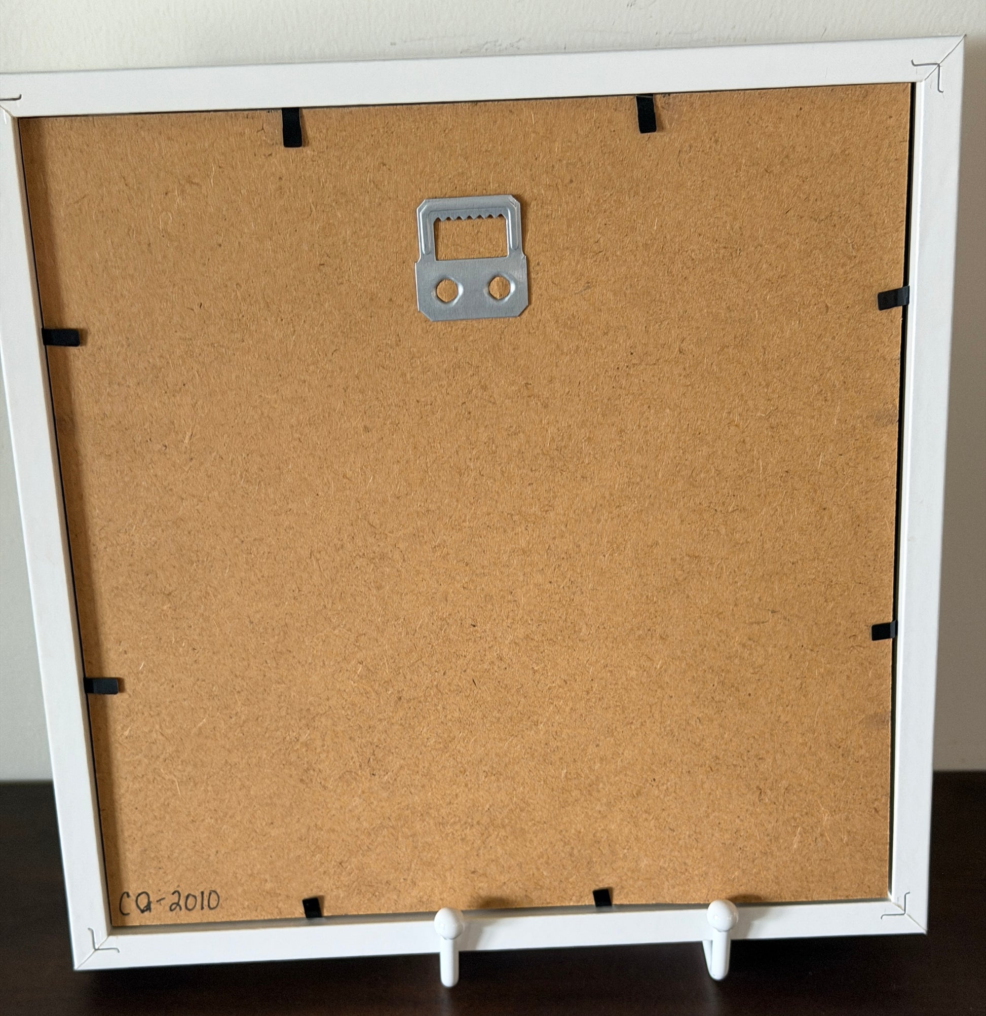 The back of a square picture frame with a white border, a brown cork backing board, a silver metal hanging loop, and white feet, shown standing on a dark surface.