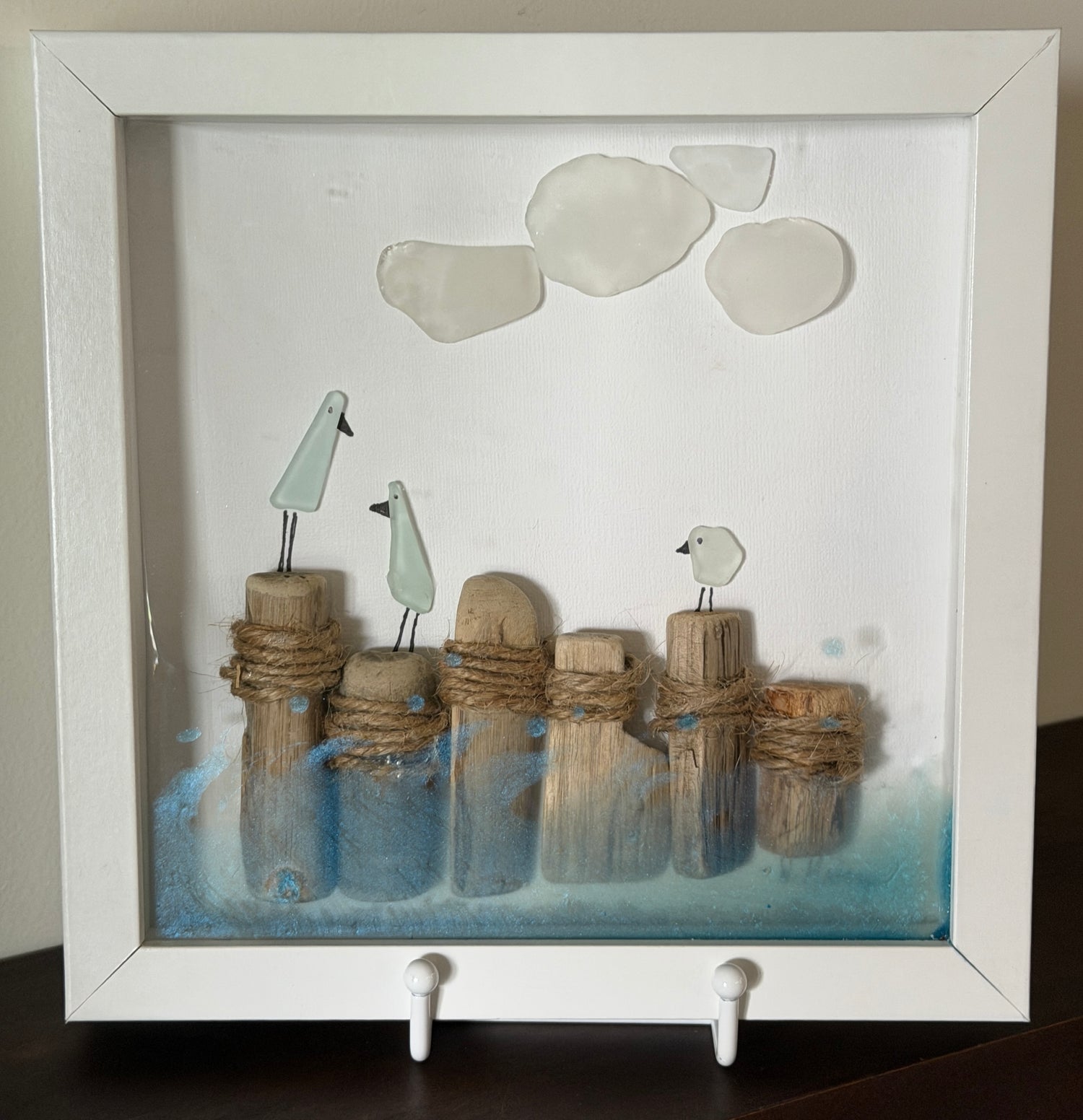 A framed shadow box art piece featuring three small white birds perched on driftwood posts wrapped in rope, with blue water-like accents at the bottom and three white sea glass stones near the top, displayed in a white frame.