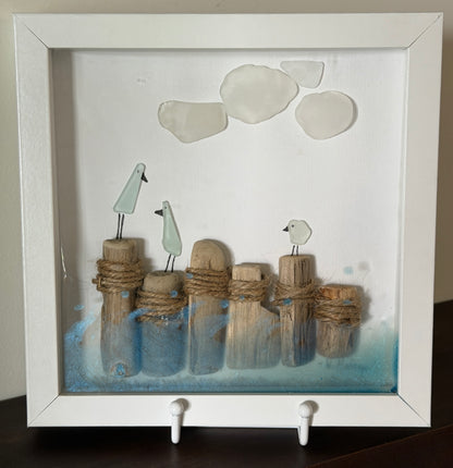 A framed shadow box art piece featuring three small white birds perched on driftwood posts wrapped in rope, with blue water-like accents at the bottom and three white sea glass stones near the top, displayed in a white frame.