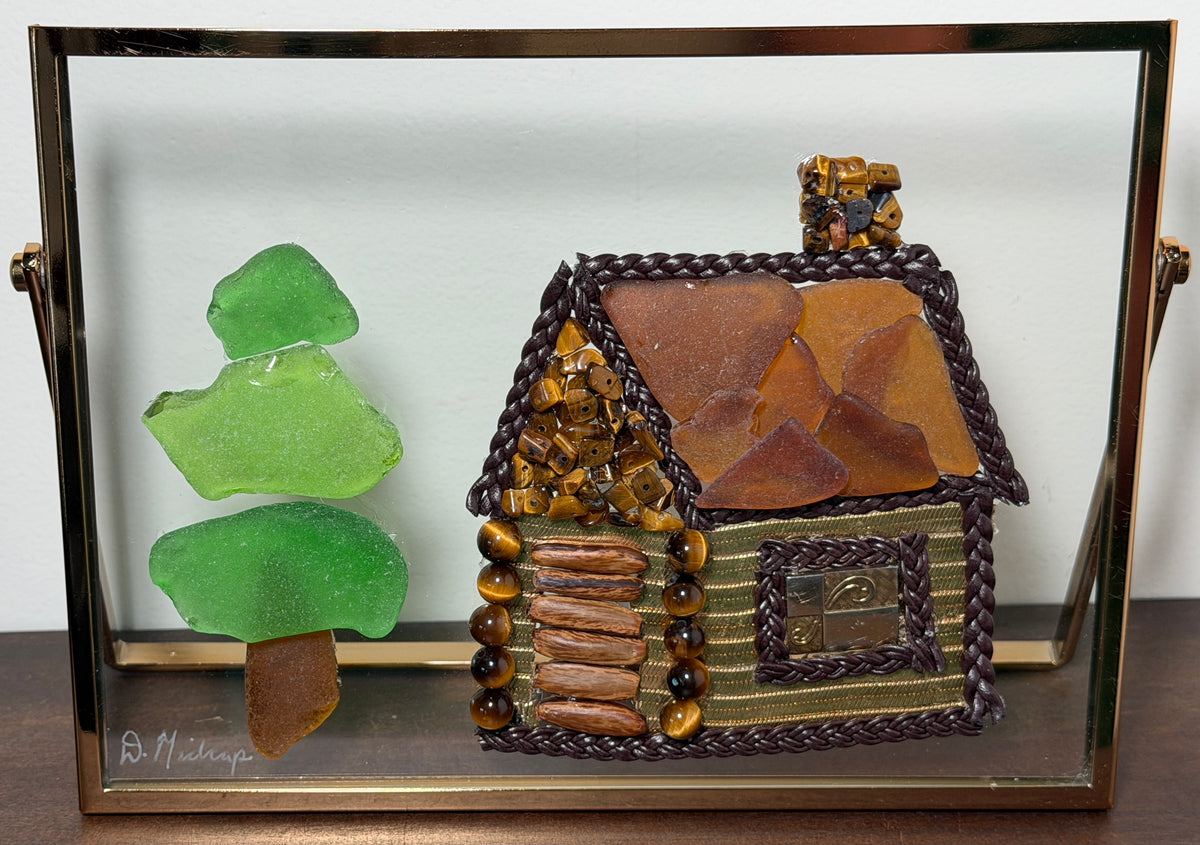 A handmade sea glass cottage and tree sculpture displayed inside a clear glass shadow box frame. The cottage is crafted from brown and tan sea glass with braided dark green rope trim and gold-toned metallic accents.
